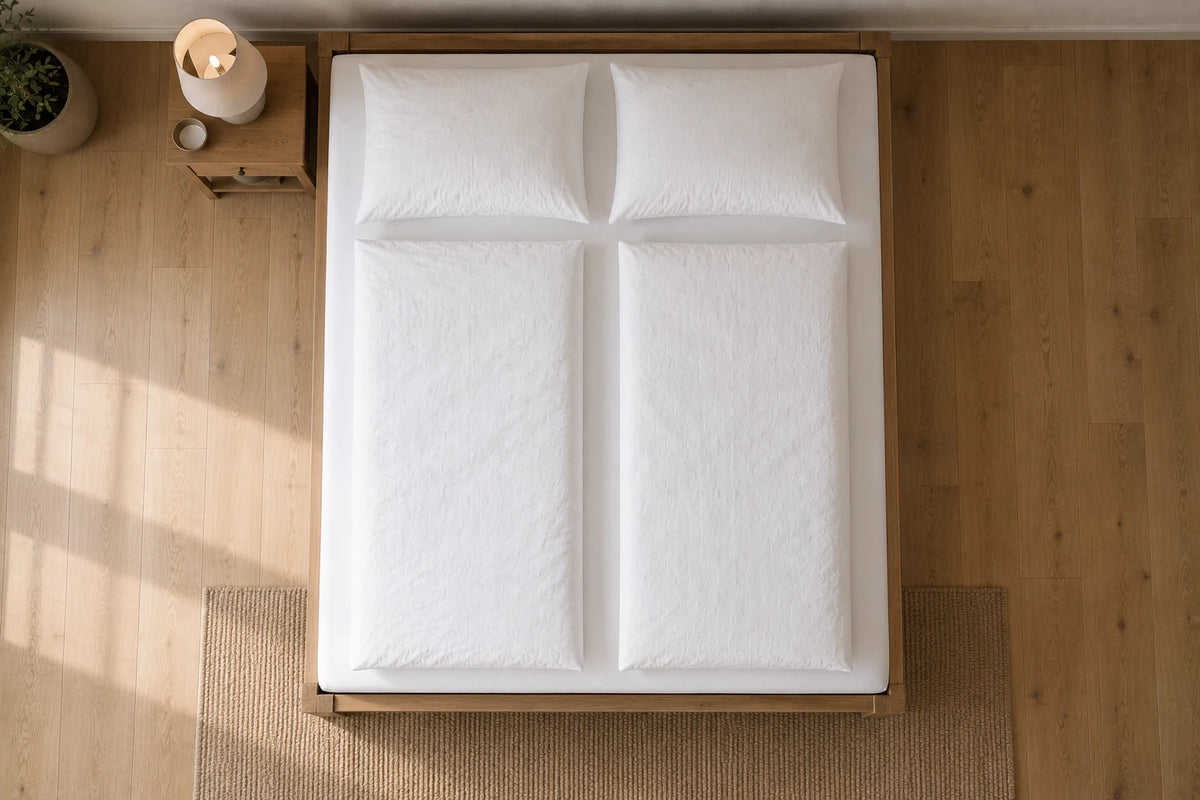 Frisch bezogenes Doppelbett von oben fotografiert, mit weißer Bettwäsche, zwei Kissen und zwei Bettdecken auf hellem Eichenboden